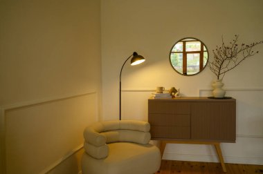 Masa lambası ile sıcak bir ışık saçan rahat dekoratif bir köşe manzarası. İç kısımda açık renkli bir koltuk, kahverengi bir çekmece, bir duvar aynası, bir vazo ve kitaplar var.