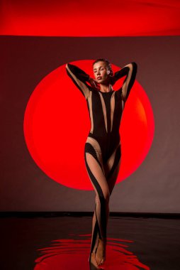 İnce, atletik bir kadın stüdyoda kırmızı bir zemine karşı poz veriyor. Stüdyo aydınlatması güneşin doğuşunu taklit eder. Genç kadın stüdyoda suyun içinde duruyor. Dar siyah bir tulum giyiyor..