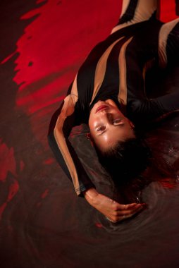 İnce, atletik bir kadın stüdyoda kırmızı bir zemine karşı poz veriyor. Stüdyo aydınlatması güneşin doğuşunu taklit eder. Genç kadın stüdyoda suyun içinde duruyor. Dar siyah bir tulum giyiyor..