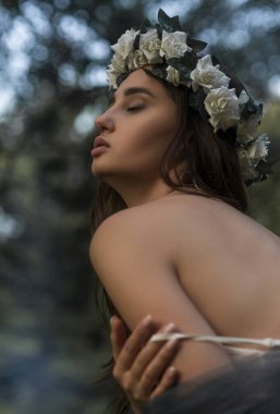 Çiçekli bir taç ve yeşil bir ormandaki su kenarında gri tül peçeli beyaz bir elbisenin ince bir modelinin samimi portresi. Hassas şefkat kavramı, doğayla duygusal bağ ve...