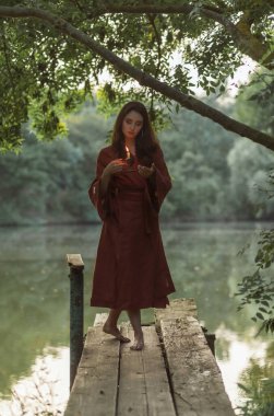 Doğal keten kıyafetli genç bir bayan, elinde yanan bir mumla kırsal ahşap bir iskelede. Yavaş yaşam, akşam meditasyonu ve yeşil orman ortamında otantik yaşam biçimi kavramı.