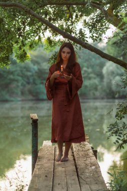 Terracotta bornozlu, elinde mumla yeşil ağaçların altında duran güzel bir kadın. Yaz ortası gündönümü kavramı, putperest gelenekleri ve deniz kenarında romantik gece aydınlatması..