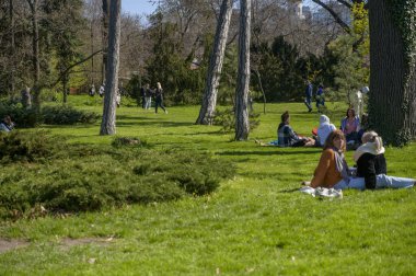 ODESSA, UKRAINE - 13 Nisan 2025: Güneşli bir bahar gününde şehir parkında yeşil çimlerin üzerinde oturan insanlar. Sosyal toplanma, açık hava rahatlama ve huzurlu şehir yaşam tarzı kavramı.