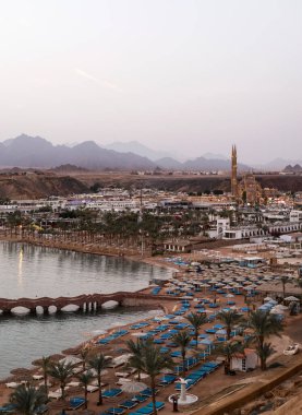 SHARM EL-SHEIKH, EGYPT - 11 Mart 2021: Alacakaranlıkta beyaz şemsiyeler ve bir köprüyle bir tatil köyünün yüksek açılı görüntüsü. Akşam keyfi ve deniz kenarında tatil konukseverliği kavramı.