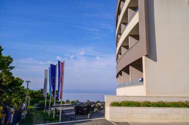 Önünde bayraklar sallanan modern bir otel ve restoranın girişi. Yeşil ağaçlar günışığı altında çağdaş mimariyi çerçeveliyorlar. Seyahat, turizm, profesyonel konukseverlik hizmeti kavramı