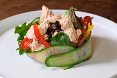 Salata, sebze, domates ve biberle servis edilen somon balığı, ince dilimlenmiş salatalığa sarılmış beyaz tabakta.