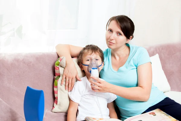 Mujer joven con hijo haciendo inhalación con un nebulizador en casa y leyendo un libro — Foto de Stock
