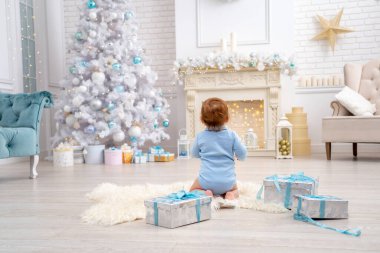 Noel süslemeli oturma odasında, arka manzaralı hediye kutularıyla kilimin üzerinde oturan sevimli bir bebek. Bir çocuk kocaman bir Noel ağacına ve ışıklara bakar. İlk yeni yıl.