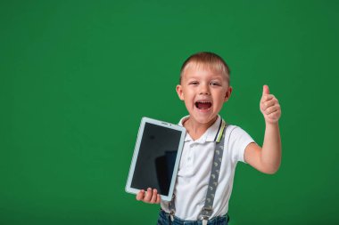 Küçük çocuk yeni bir bilgisayar tableti gösteriyor, sınıfı yeşil arka planda gösteriyor. Gülümseyen anaokulu öğrencisi, mobil bir cihazın reklamını yaparken veya çocuğun algılamasını yaparken neşeli bir ifadeyle bir cihaz tutuyor..