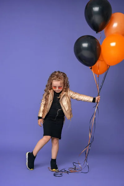 时尚的小女孩 戴着太阳镜 卷曲的头发 穿着时髦的衣服 手里拿着橙色和黑色的气球 站得很高 俯瞰着紫色的背景 — 图库照片