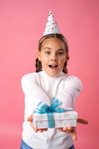 happy Birthday! Happy preteen girl in a birthday hat with a gift on a pink background. the child smiles happily and holds gifts in his hands. Advertising, discounts on toys. Copy space.