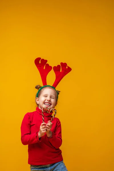 快乐的小女孩4岁在明亮的黄色背景的驯鹿鹿角 孩子很快乐 手里拿着棒棒糖 心形如初 新年销售 复制空间 — 图库照片
