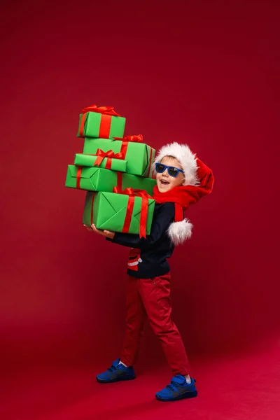 工作室里 戴着墨镜 头戴圣诞礼帽的小男孩拿着一个红色背景的圣诞礼品盒金字塔 孩子高兴地微笑着 并收到了许多圣诞礼物 — 图库照片