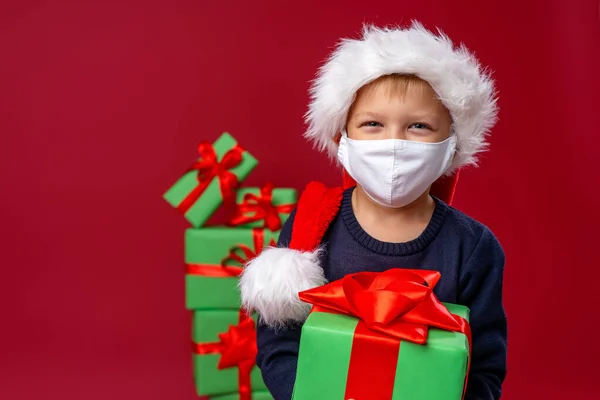little boy in a knitted hat, scarf and fluffy sweater and a medical face mask with a Christmas gift box against red background. Christmas shopping. Copy space. new normal.