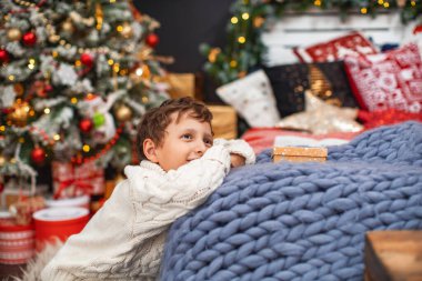 Mutlu noeller. 7 yaşındaki erkek çocuk Noel günü bir mucize hayal ediyor. Örgü bir süveter giymiş bir çocuk, Noel süslemesi için döşenmiş bir odada, örgü örülmüş bir Merino battaniyesine yaslanıyor..