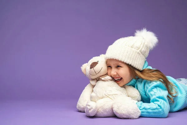 ニットセーターにブロンドの髪をした幸せな女の子4歳 帽子とミトン嘘 笑顔とスタジオで紫色の背景にテディベアを抱擁 おもちゃ 彼女の友人 冬の期間 — ストック写真
