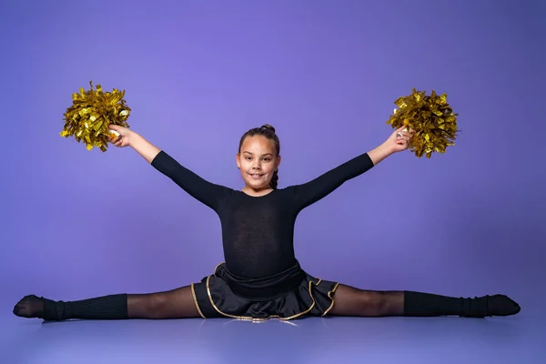 cheerleader teen girl sits on a twine in a Studio with gold POM-poms. a young gymnast does an exercise on a purple background. children\'s professional sports. Cheerleading