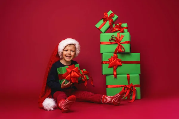 在工作室里 戴着圣诞礼帽的小男孩坐在红色背景的金字塔形圣诞礼品盒旁边 孩子很高兴看到他笑着手里拿着礼物 还收到了许多圣诞礼物 — 图库照片
