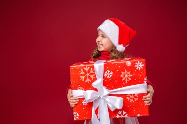 4 yaşında, parlak kırmızı arka planda Noel Baba şapkası takan mutlu küçük bir kız. Elinde bir hediye tutan komik çocuk hediye vermek için başka tarafa bakıyor. Yeni yıl satışları, reklamlar, indirimler..