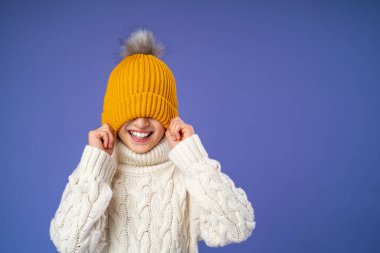 Örgü ören süveter giyen mutlu, neşeli bir çocuk mor arka planda sarı örülmüş bir şapkayı gözlerine çekiyor. Çocuk parlak kalın giysiler giydi ve kışın başlamasından mutlu. Boşluğu kopyala.