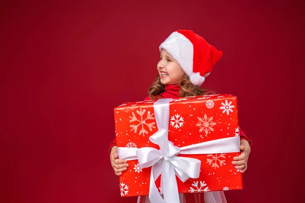 快乐的小女孩 戴着圣诞礼帽 背景鲜红 一个拿着礼物的有趣的孩子把目光移开送礼物 新年销售 — 图库照片