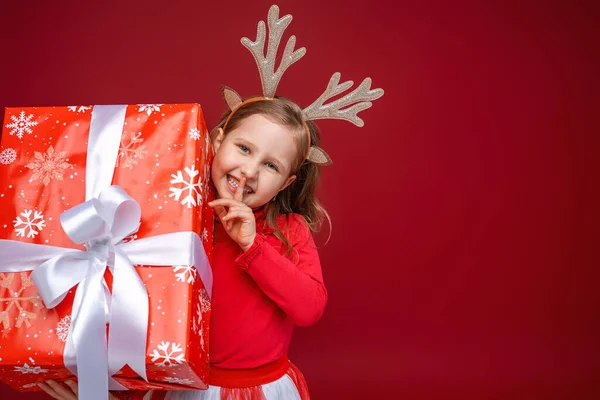 穿着红色背景鹿角的快乐小女孩 一个快乐的孩子 从盒子后面往外看 用手指捂住嘴 圣诞销售 复制空间 — 图库照片