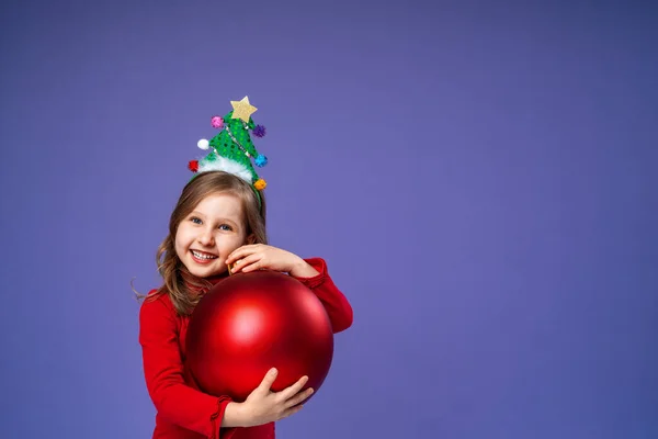 在工作室里 戴着装饰过的头巾的快乐小女孩拿着紫色背景的圣诞红球 头戴圣诞树的孩子高兴地笑着 并收到圣诞礼物 — 图库照片