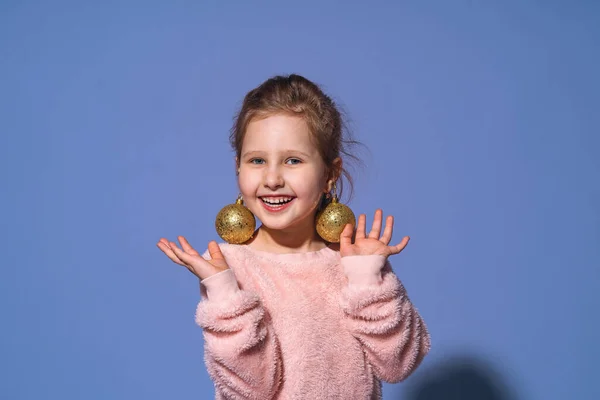 美しい魅力的な赤ちゃんの女の子は スタジオで紫色の背景にクリスマスボールアクセサリーで自分自身を飾る 幸せな女の子は彼女の耳にクリスマスボールを置き 単に笑う スペースのコピー — ストック写真