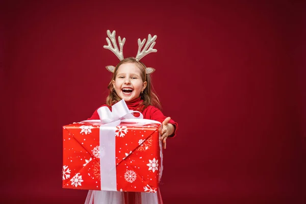 快乐的小女孩4岁在驯鹿鹿角在明亮的红色背景 孩子很高兴 手里拿着礼物 把它向前拉了拉 送礼物新年销售 — 图库照片