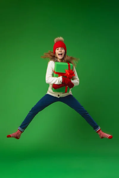 快乐的微笑的女孩 手里拿着礼品盒 头上顶着红帽 在绿色的背景上跳跃着 准备圣诞节 打折销售 动态图像 — 图库照片