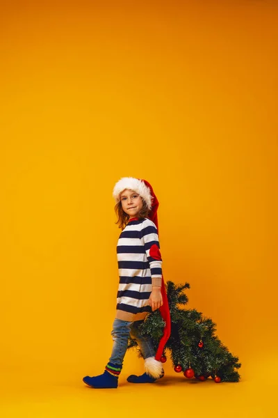 身穿条纹毛衣 头戴圣诞帽 头戴红球装饰的圣诞树的快乐小女孩 走在黄色的背景上 复制空间 — 图库照片