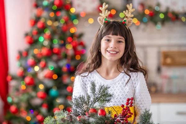 快乐的笑着的小女孩 长着鹿角和卷曲的黑头发 一个迷人的孩子穿着一件白色针织毛衣 背景是厨房 圣诞树上装饰着花环和圣诞装饰品 — 图库照片