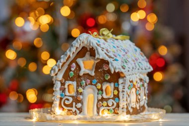 Yıldız şeklinde şekerlemelerle süslenmiş el yapımı Noel kurabiyesi evi ahşap bir masanın üzerinde duruyor. Arka planda Noel ağacı ışıkları var. Tatil için hazırlanmış lezzetli kurabiyeler..