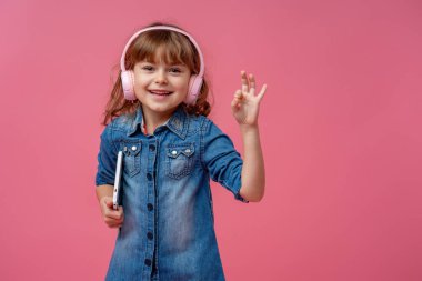 Kulaklığı ve tablet bilgisayarı olan gülümseyen küçük kız pembe arka plandaki OK sembolünü işaret ediyor. Çocuk müzik dinliyor, çalıyor ve internetten öğreniyor. Elektronik dijital öğrenme