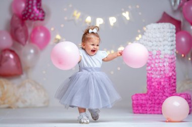 İlk yılın doğum günü. Arka planda pembe balonlar ve çelenklerle koşan beyaz elbiseli mutlu küçük kız ilk doğum gününü kutluyor. Dekorasyon doğum günü. Boşluğu kopyala.