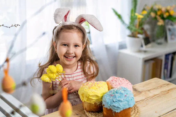 Happy Easter! funny little girl with rabbit ears sitting at the table and holding Easter cakes and eggsThe child laughs and celebrates the Easter holiday. At home, in a room decorated for the holiday