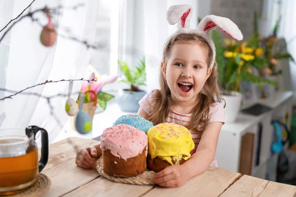 Happy Easter! funny little girl with rabbit ears sitting at the table and holding Easter cakes. The child laughs and celebrates the Easter holiday. At home, in a room decorated for the holiday