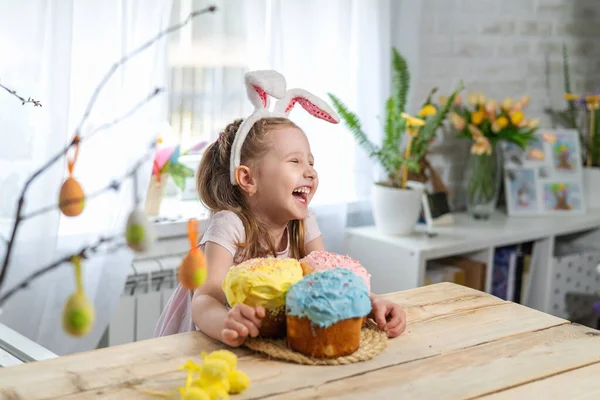 Happy Easter! funny little girl with rabbit ears sitting at the table and holding Easter cakes. The child laughs and celebrates the Easter holiday. At home, in a room decorated for the holiday