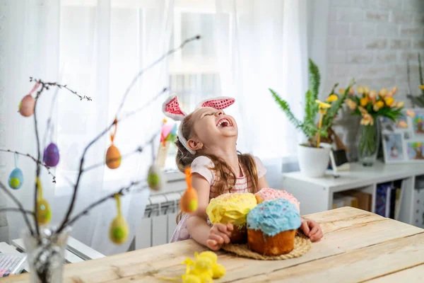 Happy Easter! funny little girl with rabbit ears sitting at the table and holding Easter cakes. The child laughs and celebrates the Easter holiday. At home, in a room decorated for the holiday