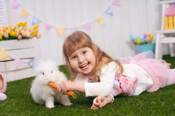 부활절 날귀여운 아기의 소녀가 색깔있는 달걀을 가지고 토끼에게 당근을 먹인다 — 스톡 사진