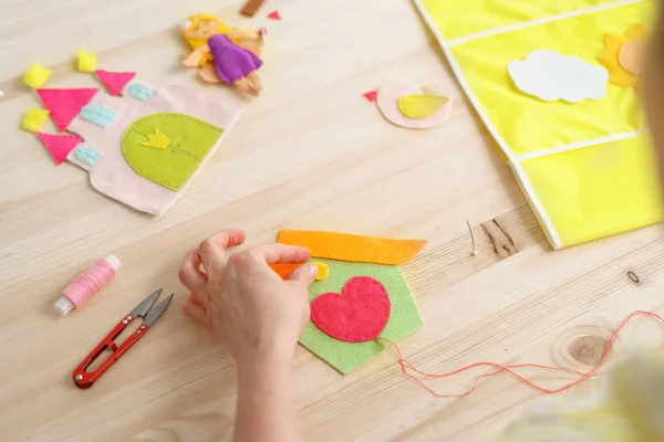 Close-up women's hands doing crafts, sewing house with heart made of felt. Accessories for needlework on wooden table. Spring or summer crafts for children and adults. Creative DIY for room decoration