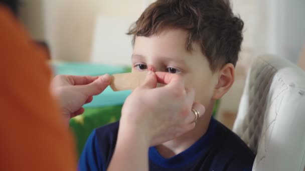 Een vrouw plakt een pleister op de wond van een kind. de jongen die zijn gezicht verwondde krijgt medische zorg thuis. diepe wond op de neus en blauwe plekken met krassen op het gezicht. — Stockvideo