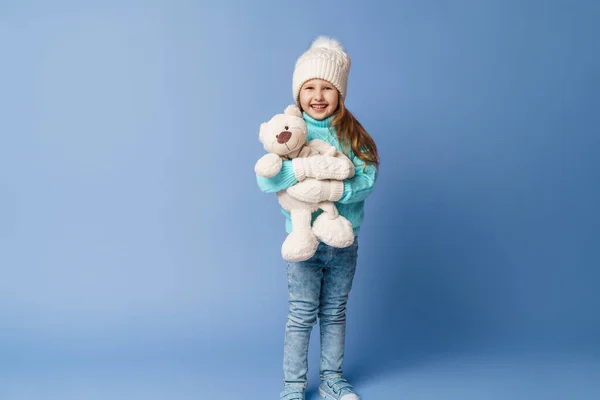 快乐的5岁小女孩 金发碧眼 头戴针织毛衣帽 头戴手套 笑着抱着一只泰迪熊在工作室的蓝色背景上 这个玩具是她的朋友 冬天的时候假日周末 — 图库照片