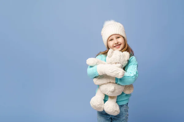 ニットのセーターキャップにブロンドの髪をした幸せな5歳の女の子とミトン スタジオの青い背景にテディベアを抱いています おもちゃは彼女の友人です 冬時間だ — ストック写真