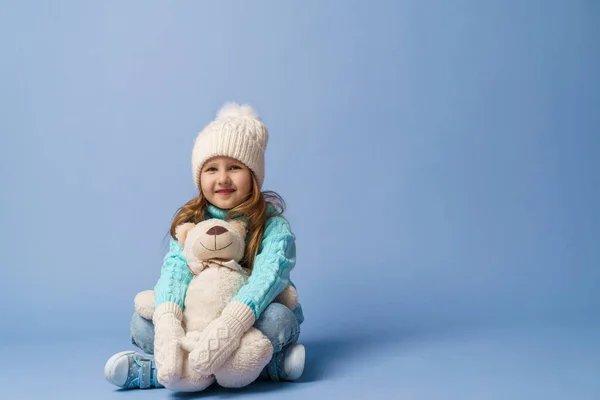 幸せな小さな5歳の女の子とともにブロンド髪でニットセーターキャップとミトン 笑顔と抱擁テディベアはスタジオで青の背景に座っています おもちゃは彼女の友人だ 冬時間だ — ストック写真