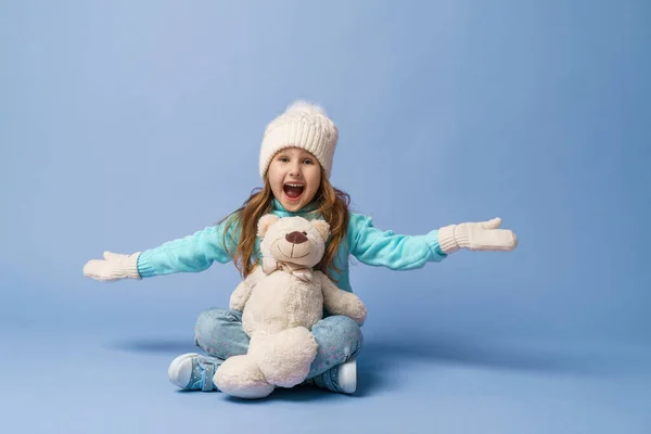 幸せな小さな5歳の女の子とともにブロンド髪でニットセーターキャップとミトン 笑顔と抱擁テディベアはスタジオで青の背景に座っています おもちゃは彼女の友人だ 冬時間だ — ストック写真