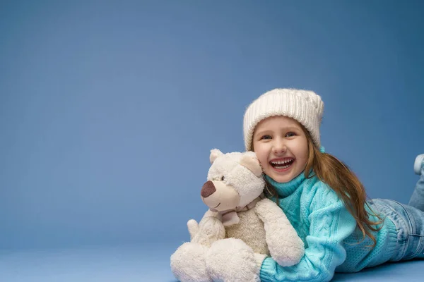 快乐的5岁小女孩 金发碧眼 头戴针织毛衣帽 头戴手套 笑着抱着泰迪熊躺在工作室的蓝色背景上 玩具是她的朋友 冬天的时候假日周末 — 图库照片