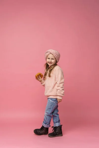 穿着毛绒绒毛衣 头戴头巾的小女孩 手里拿着甜甜圈 微笑着 甜开胃菜 好吃的糕点 一个孩子在一个全长的工作室里 在粉色的背景上展示了一份美味的甜点 — 图库照片
