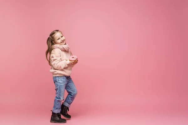 ふわふわのセーターとヘッドバンドの女の子がドーナツを手に笑っていました 甘い前菜 おいしいペストリー 子供はピンクの背景においしいデザートを見せてくれます — ストック写真