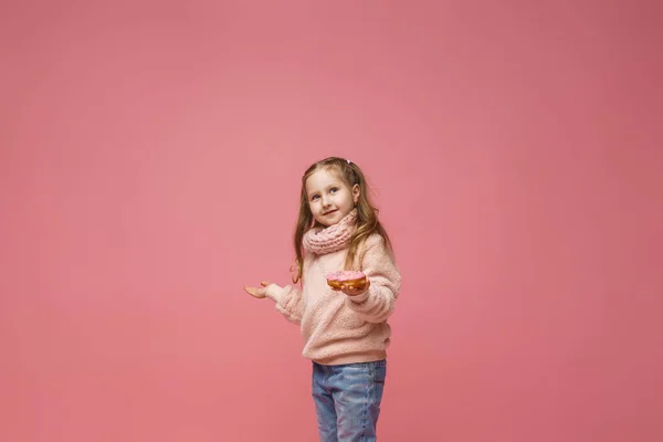 穿着毛绒绒毛衣 头戴头巾的小女孩 手里拿着甜甜圈 微笑着 甜开胃菜 好吃的糕点 在演播室里 一个孩子在粉色背景上展示了一份美味的甜点 — 图库照片